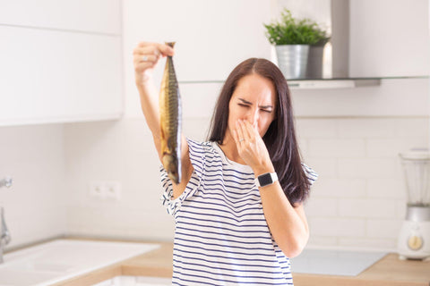 Woman disgusted from a smell of a fish she holds in hand holds her nose standing in the kitchen at home