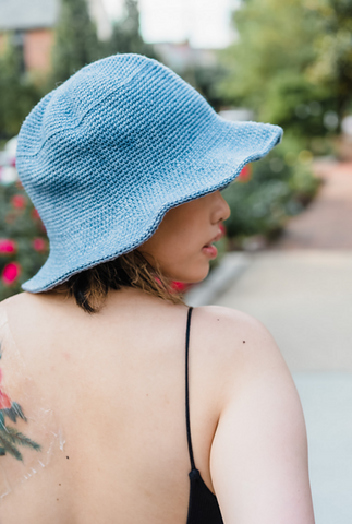 a  woman shot from behind wearing a blue bucket hat, there is greenery in the background.