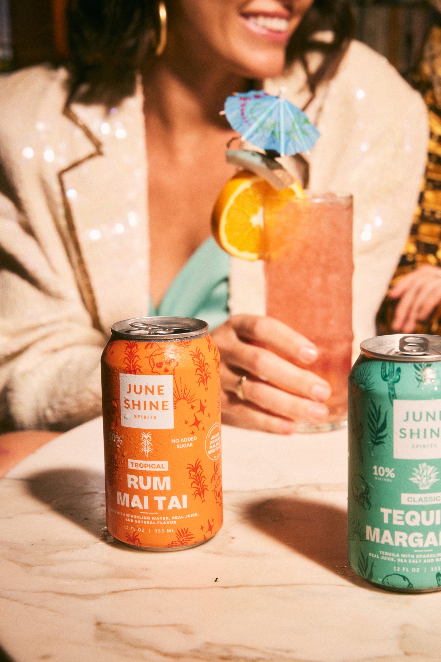 JuneShine Spirits cans on bar table with woman in the background holding a cocktail glass