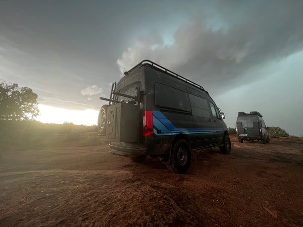 Two Mercedes Benz Revel Adventure Vans are sitting on a dirt road after a heavy rain.  There are dark skies mixed with blue and the bright sun is a ribbon of yellow light at the horizon.