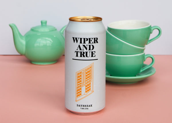 A white can with an orange illustration of a window is on a pale pink flat lay. Three teal piled tea cups are on a saucer and a teapot of the same colour is in the background against a white backdrop.