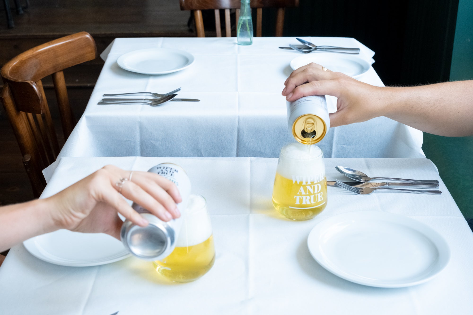 Two hands pouring beer from white cans into glasses on a table covered with a white tablecloth