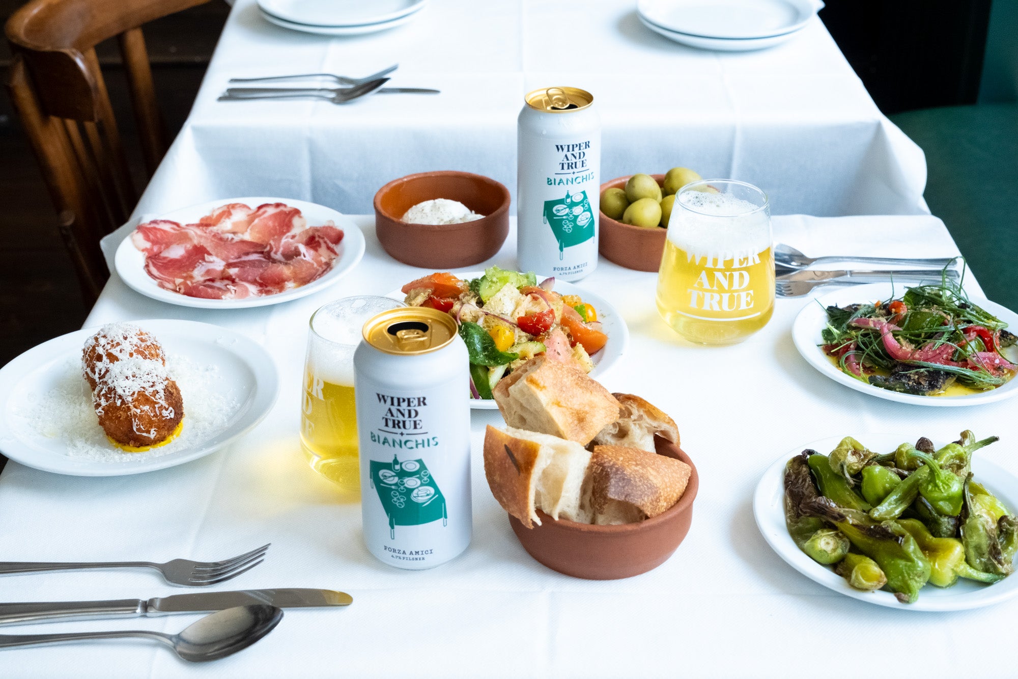 A table with a white table cloth, full of small plates of food and two beers