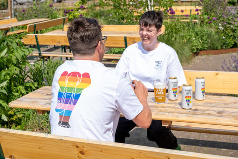 Two light-skinned people with short brown hair sit opposite each other at a wooden table. They both wear white t-shirts with a rainbow elephant on the back.