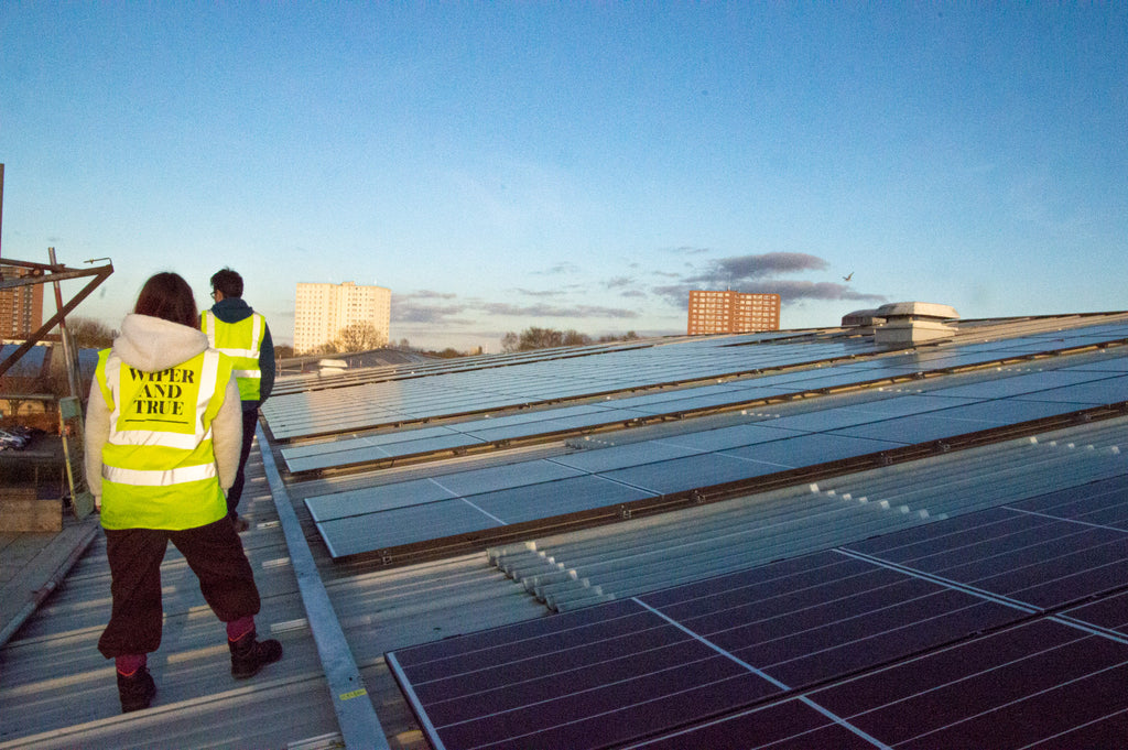 Two people stand next to solar panels on a roof. The sky behind is blue without a cloud.