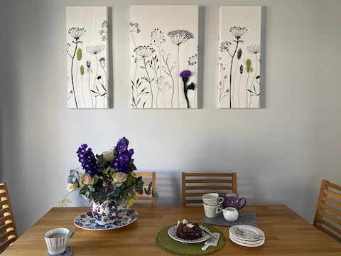 A photo of a dining table with purple floral, Emma Bridgewater vase filled with fresh co-ordinating flowers. The table is set with a tea pot, tea cups and chocolate cake. Three canvases are on the wall. These have black and white wild flower scenes on them, each canvas has one flower embellished and made 3D in purple or green using felt, wool and 3D plastic.