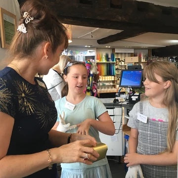 Photo of two girls being taught how to wrap ribbon around a chocolate box by a female member of the shop team.
