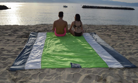 A couple sittiing on a green ECCOSOPHY beach blanket at the beach and watching the sunset.