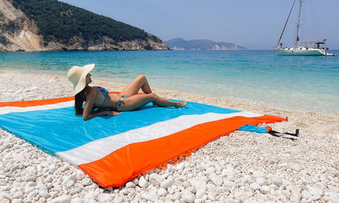 lady laying on a blue and orange ECCOSOPHY beach blanket. She is sitting on white pebbles looking into the beautiful blue sea.