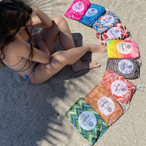 Girl on a sandy beach looking at a collection of sand free, quick dry microfiber towels made by ECCOSOPHY. The microfiber towels are in their travel pouch. All ECCOSOPHY beach towels are double sided and have vibrant colors.