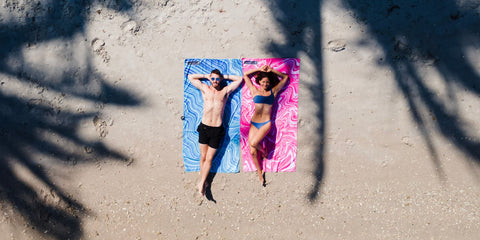 A man laying on a sandy beach on an ECCOSOPHY blue sand free beach towel and a woman laying next to him on a pink ECCOSOPHY quick dry towel.