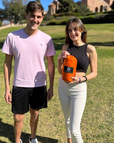two college students on campus holding a compact and lightweight Beach blanket in it's travel pouch. This pictures shows how a Large sized ECCOSOPHY beach blanket can fold down small into its travel pouch.