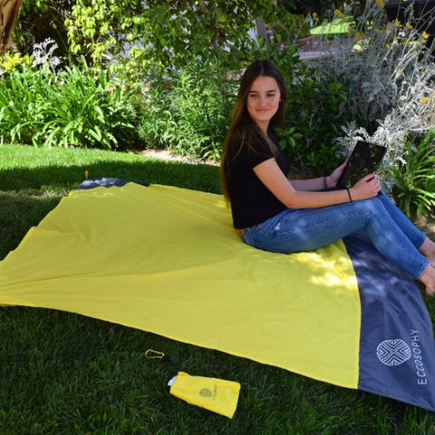 A college student sitting on campus on a waterproof beach blanket made by ECCOSOPHY. The student is holding a computer and working on the lawn.