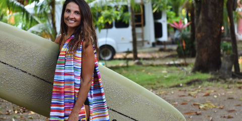 A young lady holding a surf board that has a Muti colored stripped microfiber beach towel from ECCOSOPHY thrown over her shoulder. The ECCOSOPHY surf towel is called Key West.