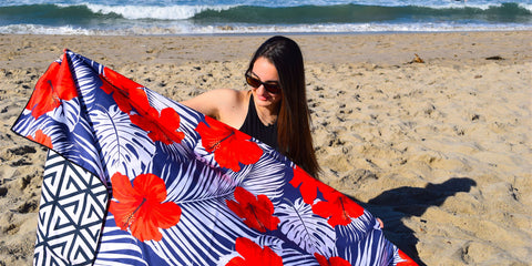 A young woman on a sandy beach holding up a red, white and blue sand proof beach towel from ECCOSOPHY. This sand free microfiber towel is designed to fold down small and is travel friendly. 