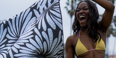A young black lady who is laughing and holding a black and white ECCOSOPHY microfiber beach towel. This towel is called Lovina after the beach in Bali. She is laughing because she is excited about the soundproof and quick drying qualities of ECCOSOPHY'S beach towels.