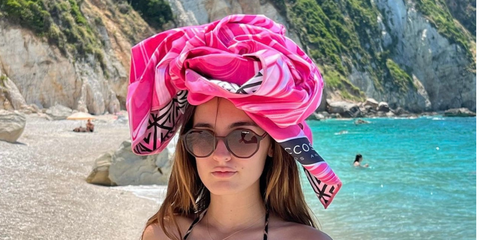A young woman on a beautiful blue beach with an ECCOSOPHY pink microfiber beach towel on her head.