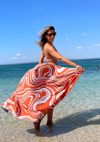 A lady on the beach with her feet in the water holding an ECCOSOPHY eco friendly microfiber beach towel around her waist. The beach towel is flowing behind her in the wind.