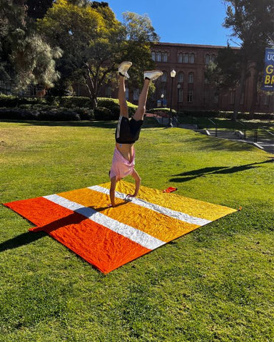 A college student on a university campus doing a handstand on an ECCOSOPHY Beach blanket. The blanket is three shades of orange.