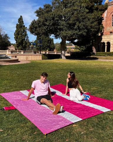 two college students on campus sitting on an ECCOSOPHY XL beach blanket. The beach blanket is three shades of pink.