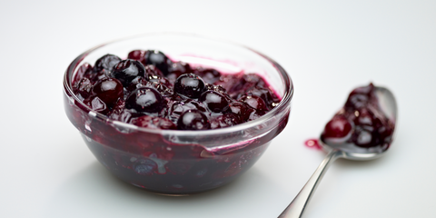 blueberry compote sits in a small glass bowl. there is a spoon beside the bowl with some of the compote on it