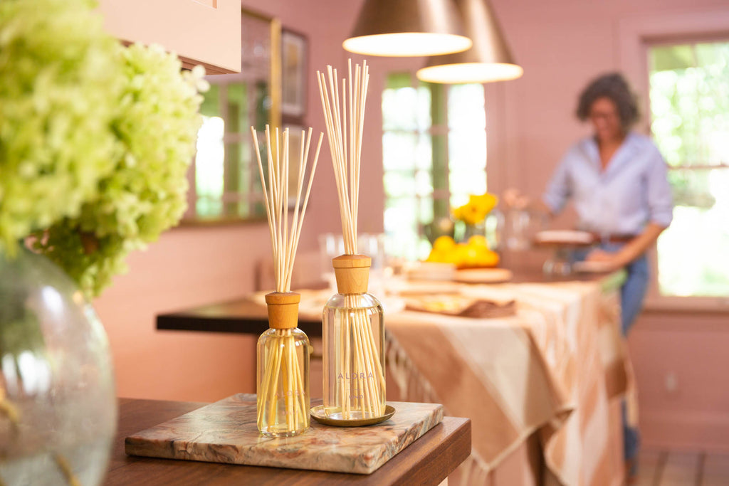 One small and one large reed diffuser in a pink kitchen with woman in back cutting a cake