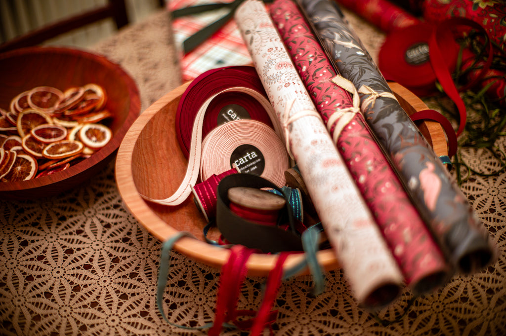 Large spools of Carta ribbons laying next to patterned wrapping paper rolls and a bowl of dried orange slices