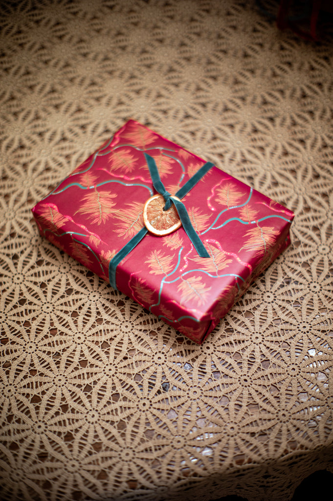 Gift wrapped in red botanical wrapping paper and tied with a thin green velvet ribbon tied with a dried orange slice