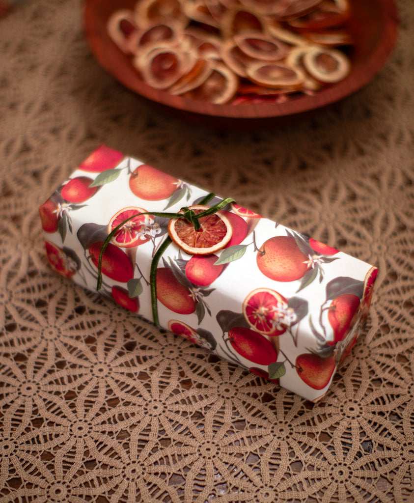 Rectangular box wrapped in orange-printed wrapping paper, tied with a thin green ribbon with a dried orange slice tied on