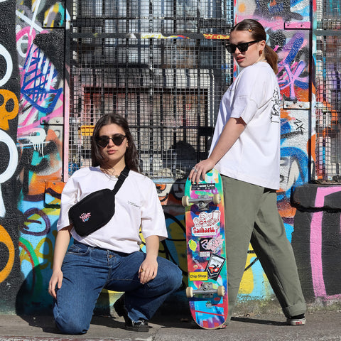 Two people standing in front of a graffiti'd wall, one holding a skateboard