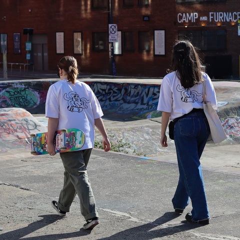 Two people holding skate boards walking through a skate park with white t-shirts