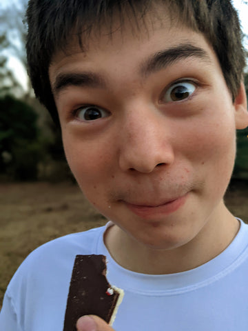 A boy with short brown hair makes a wide eyed and excited face as he chomps on a piece of Xocolatl's peppermint bark.