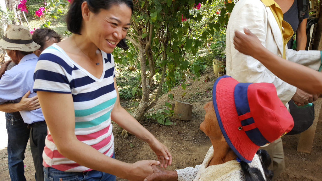 A woman with short brown hair, wearing a white shirt with blue, teal, pink stripes, stands next to an older woman who is sitting down wearing a tan shirt and red bucket hat with blue band across it. Both women are smiling, but the sitting woman's face is partially obscured.