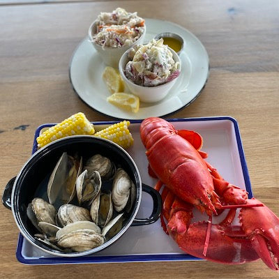 A whole steamed lobster, corn, and a pot of steamed clams on a tray next to a plate with slaw, melted butter, and potato salad 