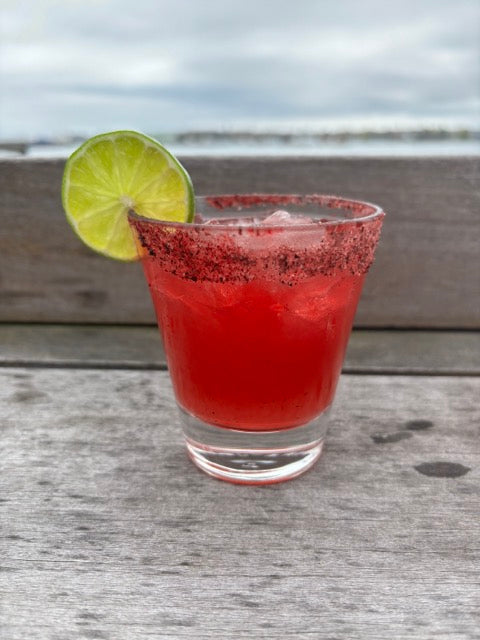 a red cocktail with a wedge of lime and a salted rim on the deck in front of the ocean