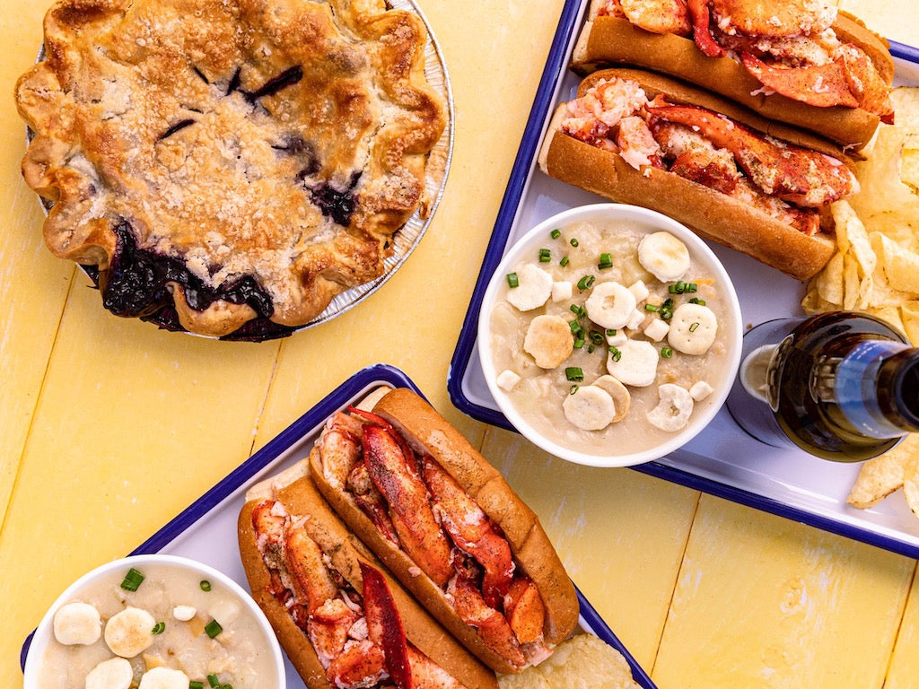 Lobster rolls, clam chowder, kettle chips, and blueberry soda on white trays next to a blueberry pie