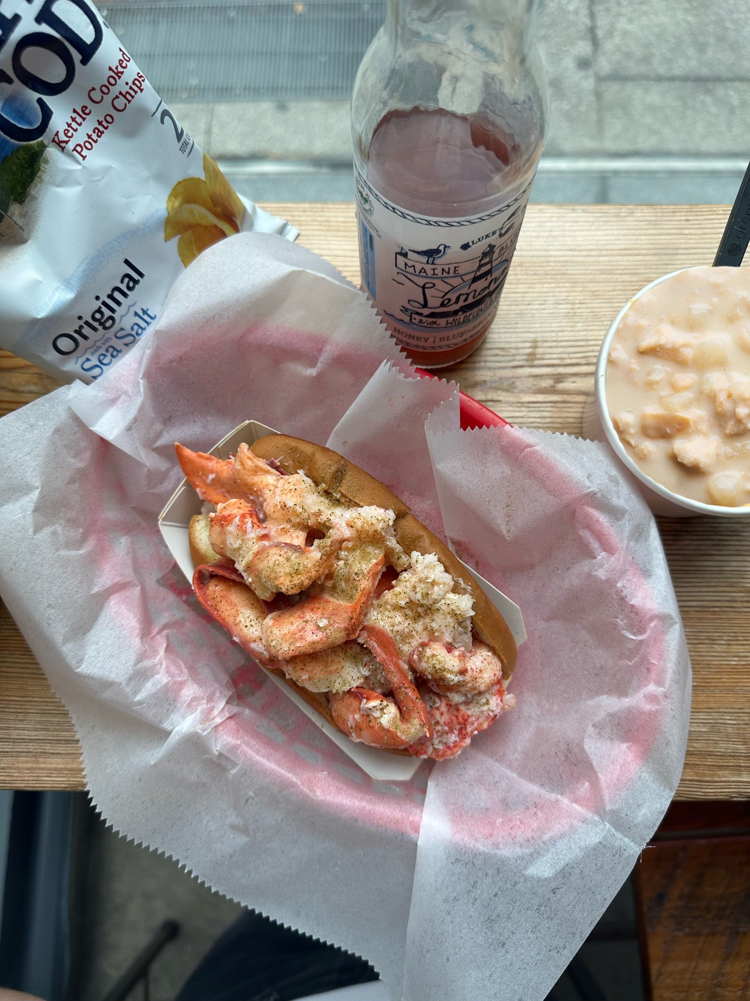 A lobster roll in a red basket lined with wax paper next to a bag of chips, a cup of chowder, and a blueberry lemonade
