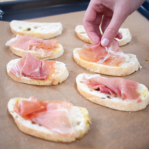 Wrijf de gegrilde broodjes licht in met knoflook en leg ze op een schaal. Halveer ondertussen de plakken ham in de breedte en verdeel ze over de bruschette.