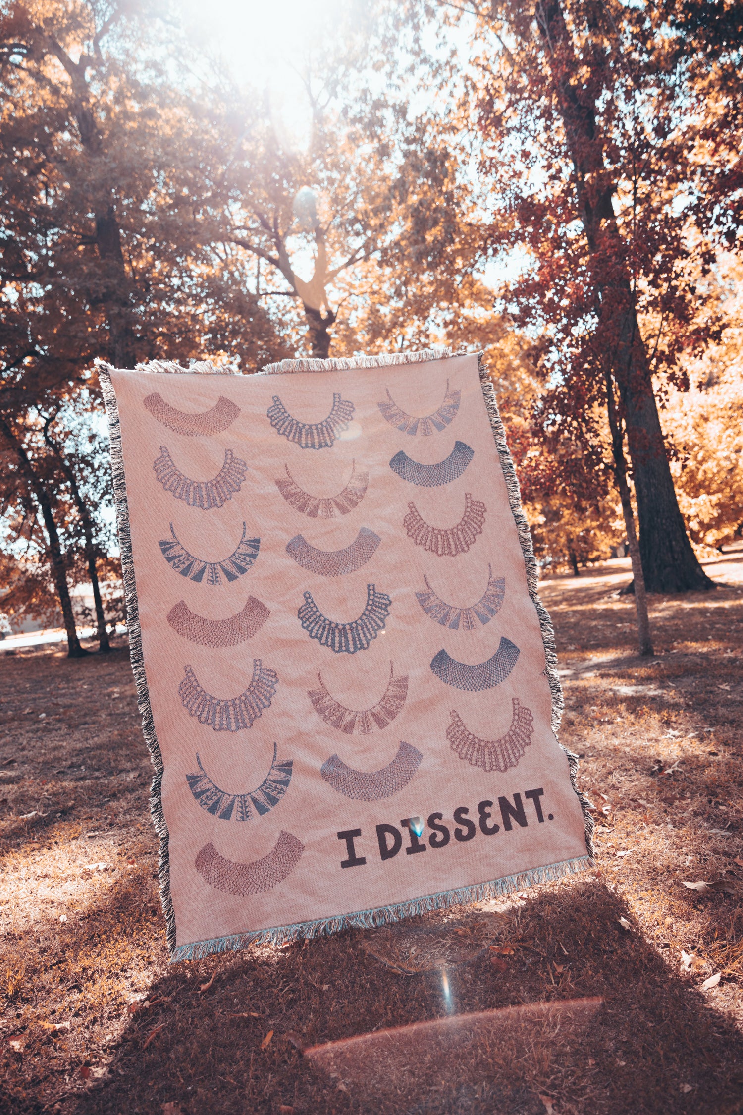 I Dissent lettering beneath pattern of Ruth Bader Ginsburg dissent collars on woven blanket. Hues of pink and purple on light pink background.