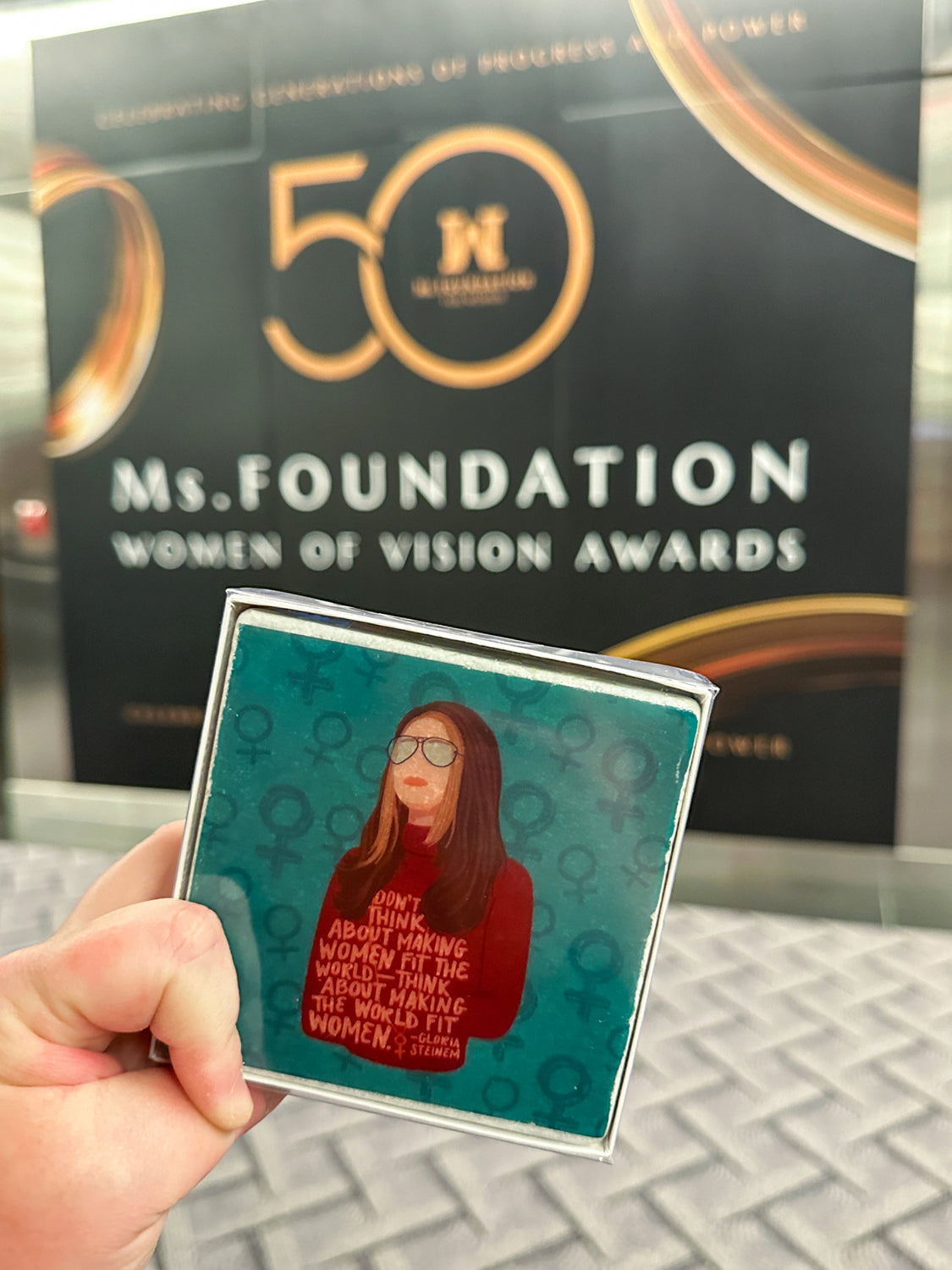 Gloria Steinem Coasters held up in front of 50th anniversary gala sign