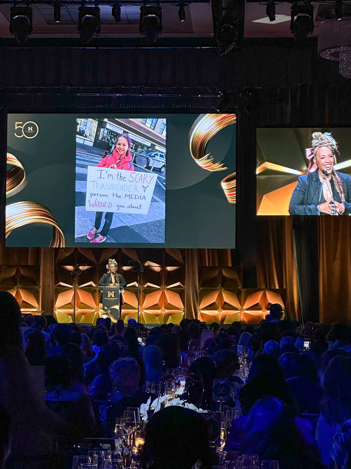 Black woman speaking at a gala with photo of young transgender girl holding poster on the screen behind her