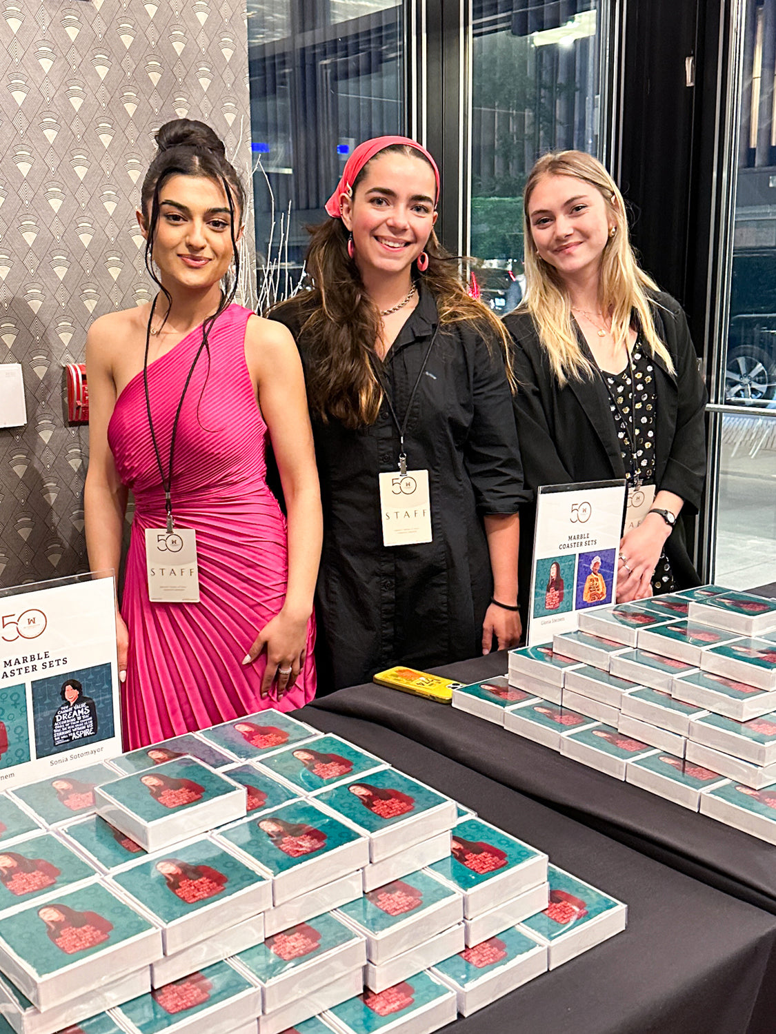 three young women standing behind two tables filled with gloria steinem coasters