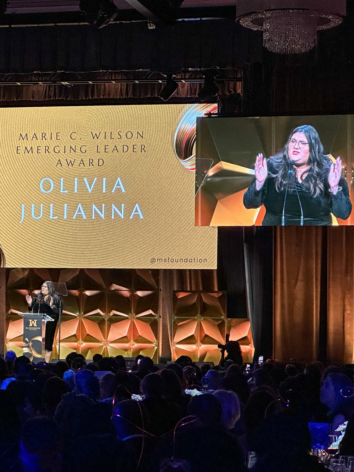 olivia julianna, woman in black dress with her hands up speaking on stage at a gala