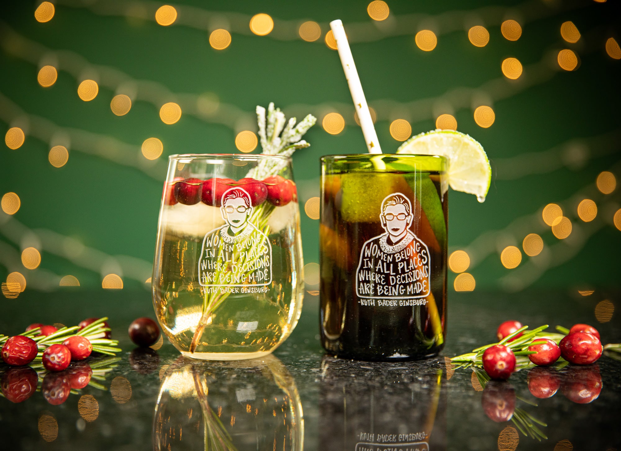 Ruth Bader Ginsburg illustration on barware; clear stemless wine glass and wine glass tumbler made from recycled green glass wine bottle; holiday themed drinks in both glasses with blurred christmas lights behind them.