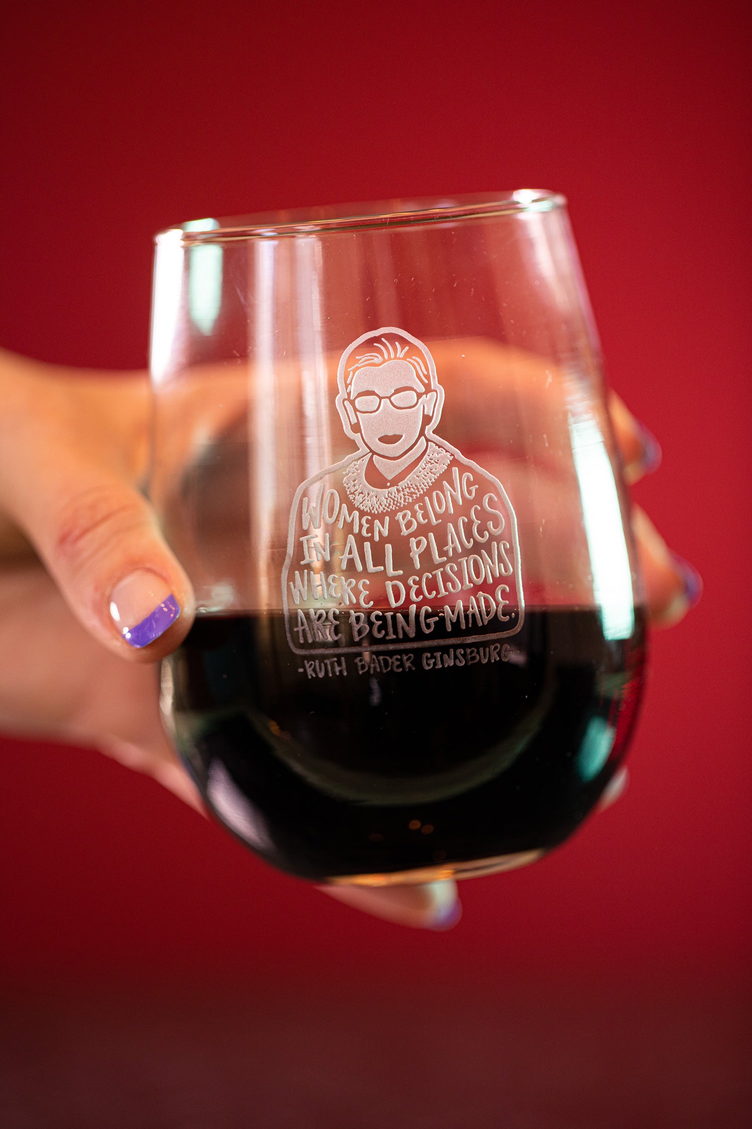 Woman holding stemless wine glass featuring sand carved illustration of Ruth Bader Ginsburg; glass filled with red wine on red background