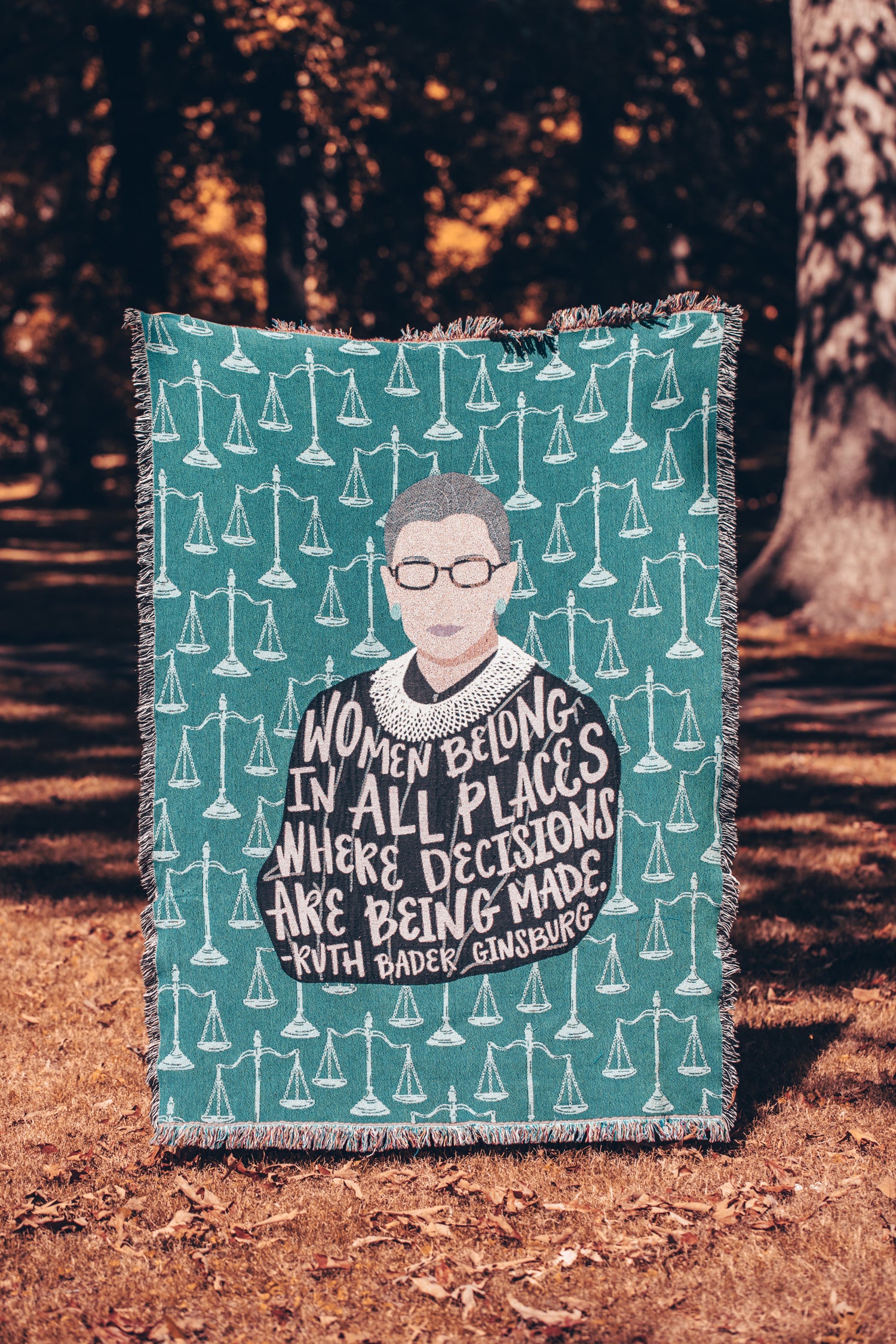 Fringe edged woven blanket tapestry featuring illustration of Ruth Bader Ginsburg with quote "Women belong in all places where decisions are being made." Includes pattern of justice scales on teal background. Blanket pictured being held up to full size in park during fall.