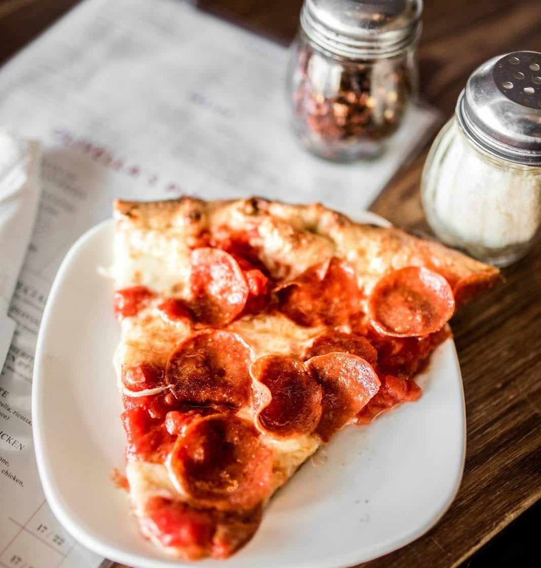 ALT IMG TXT: A slice of pepperoni pizza on a white plate atop a table with jars of seasoning nearby.