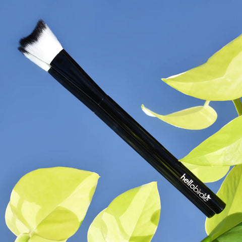 A Hello Birdie Nail and Lash Lab black lash cleansing brush with white and black curved bristles resting on a mirrored blue sky background with green leaves around