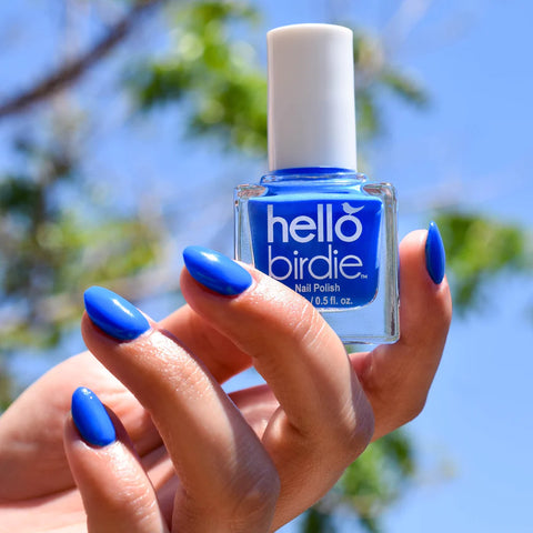 A model's hand holding a square bottle with white round lid. The bottle reads Hello Birdie, and the technicolor blue polish is seen through the clear glass bottle. The background is a blue sky and tree.
