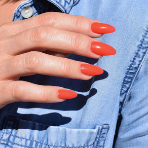 Gel polish hand-painted press on nails on one had. The almond shaped nails are in a vibrant and bright orange with a hi-gloss finish. The models hand rests over a denim shirt.